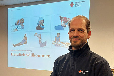 2025-11-24_Sebastian_Zuber_660x440px.jpg Der Ausbilder steht im Unterrichtsraum und blickt freundlich in die Kamera. Hinter ihm ist eine Folie aus der Ausbildung an die Wand projiziert. Herzlich willkommen steht darauf.